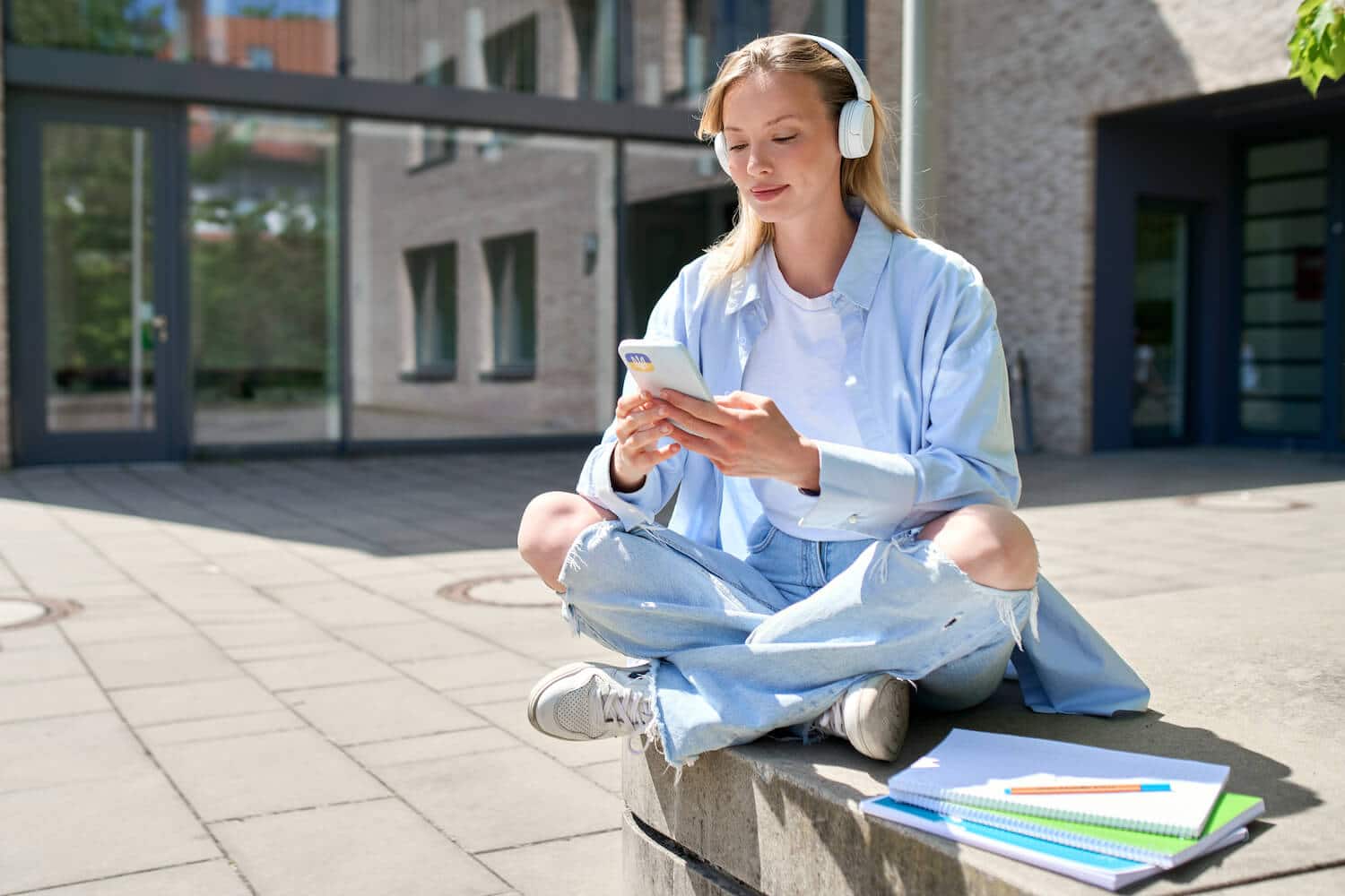Eine junge Frau sitzt vor ihrer Schule und hört während ihrer Mittagspause eine Podcast Episode.